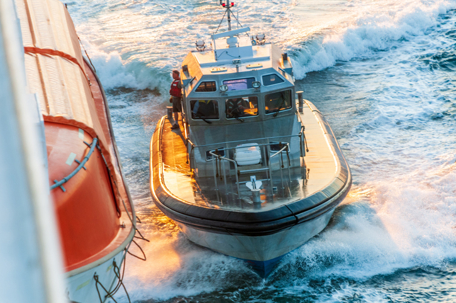 Rescue boat speeding beside a larger vessel on rough water, with waves splashing and crew visible on deck during response.