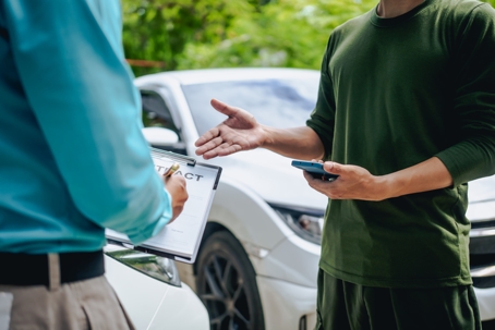 Driver at roadside car accident scene speaking with insurance adjuster holding clipboard beside damaged vehicle.