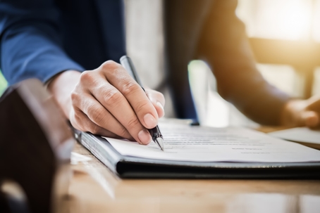A lawyer in a suit signing a legal document on a clipboard at a desk, with natural sunlight illuminating the workspace.