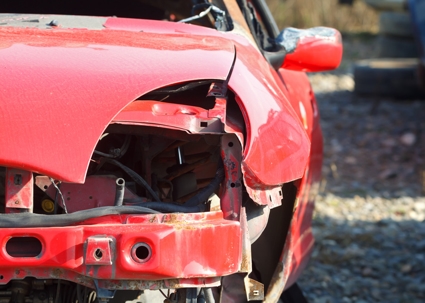 A red car with severe front-end damage, missing headlight and bumper, parked on gravel after a collision.
