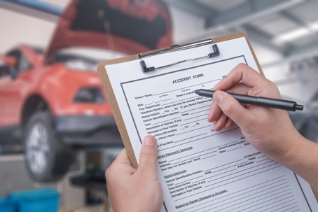 Hands holding a clipboard with a car accident report form being filled out, with a damaged car visible in the background.