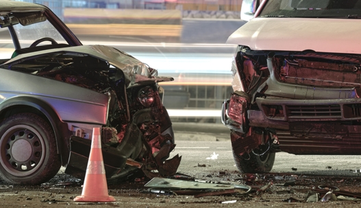 Two cars with severe front-end damage after a nighttime collision, with a traffic cone placed between them.