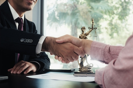 Two people shaking hands over legal documents with a Lady Justice statue in the background, suggesting a legal agreement.