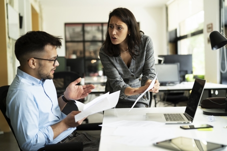 A woman angrily confronts a seated male coworker in an office, holding documents as they argue at a cluttered desk.