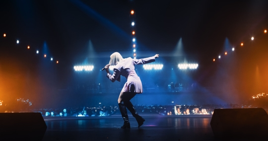 A singer performs on a brightly lit concert stage, facing a large audience with colorful spotlights glowing behind her.