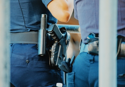 Two police officers in uniform, shown from the waist down, with holstered firearms and batons on their duty belts.