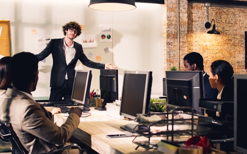A man in a suit presents data to colleagues in a modern office, standing near a whiteboard with charts and graphs.