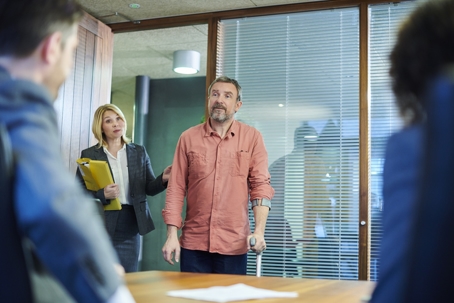 A man in a salmon shirt stands and speaks in a meeting room while a woman beside him holds folders and looks at seated colleagues.