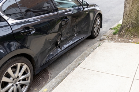 A black sedan with significant damage to the driver's side door, parked on a city street next to a curb and sidewalk.