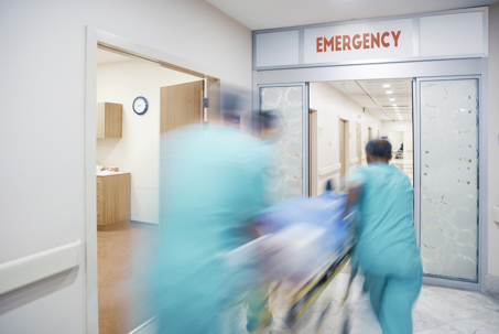 Medical staff rush a patient on a stretcher through hospital emergency room doors marked “Emergency.”