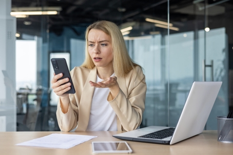 Woman in an office looks confused while checking her smartphone, with a laptop and documents on the desk.