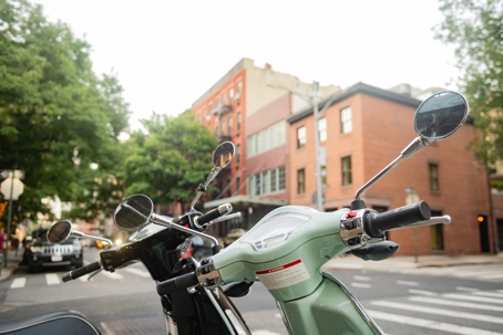 Two parked mopeds with side mirrors in focus on a quiet city street, with brick buildings and trees in the background.