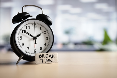 Classic black alarm clock showing 10:10 on a desk with a small wooden sign that reads “Break Time!” in an office setting.