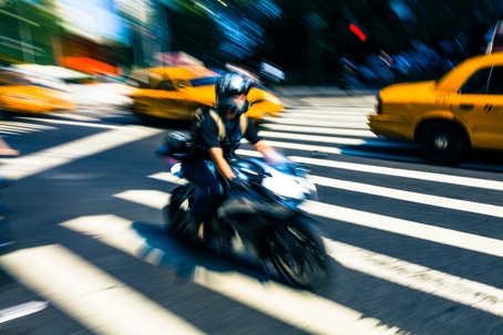 Motorcyclist speeding through crosswalk in New York City traffic, blurring past yellow taxis—risk of collision appears high.