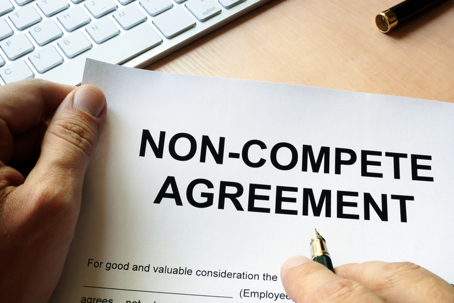 Close-up of hands holding a document titled “Non-Compete Agreement” on a desk with keyboard and pen, showing an employment contract.