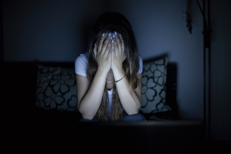 A woman sitting in a dark room covers her face with her hands, appearing distressed or overwhelmed.