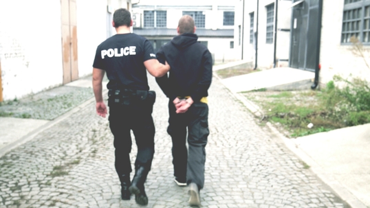 A police officer escorts a handcuffed man down a cobblestone alleyway near industrial-style buildings during daylight.