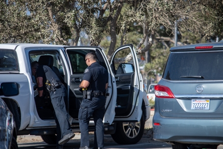 Two police officers inspect a white pickup truck with doors open beside parked cars in a lot.
