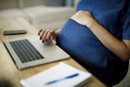 Pregnant woman working on laptop at desk, one hand on belly, phone and notebook nearby, symbolizing pregnancy rights.