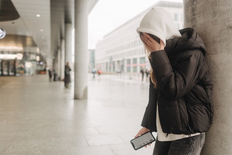 A person in a hoodie and jacket leans against a concrete pillar in a public space, covering their face while holding a smartphone.