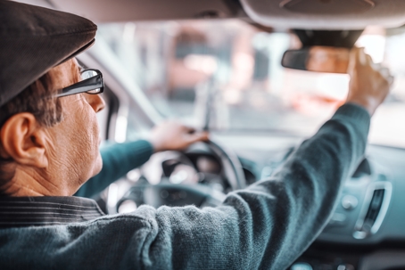 Senior driver wearing glasses adjusts the rearview mirror while seated behind the wheel of a car, viewed from inside the vehicle.