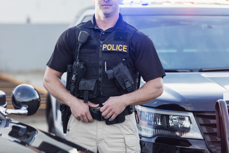 Police officer in tactical vest standing in front of a patrol car with flashing lights, holding gear at waist during duty scene