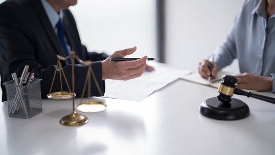 Two people in formal attire discuss legal documents at a desk with a gavel and justice scales, suggesting a legal consultation.