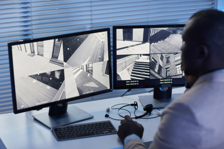 Security staff monitoring multiple CCTV camera feeds on dual monitors showing building entrances, hallways, and stairwells.