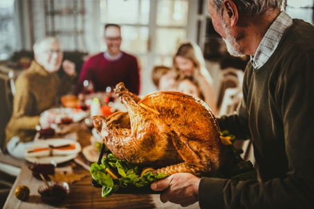 A family gathered around a dining table as an older man serves a roasted turkey at a warm, festive holiday meal.