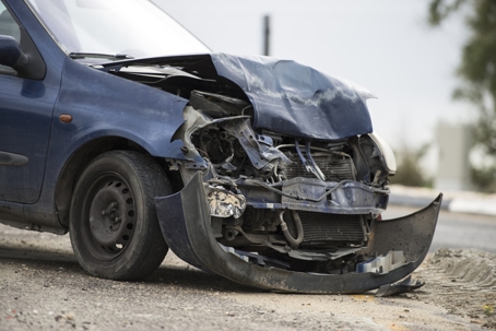 A damaged blue car with a severely crumpled front end and detached bumper sits on the side of the road after a collision.