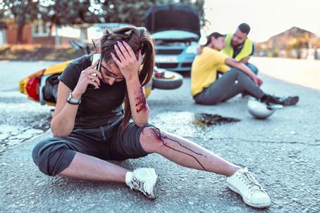 Injured woman with bleeding cuts calls for help after road accident, while another victim is assisted near a crashed car and bike.