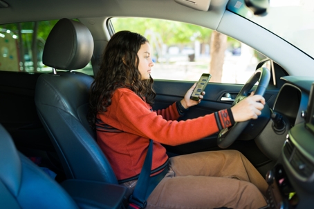 A teenage driver looking at her phone while holding the steering wheel inside a parked car.